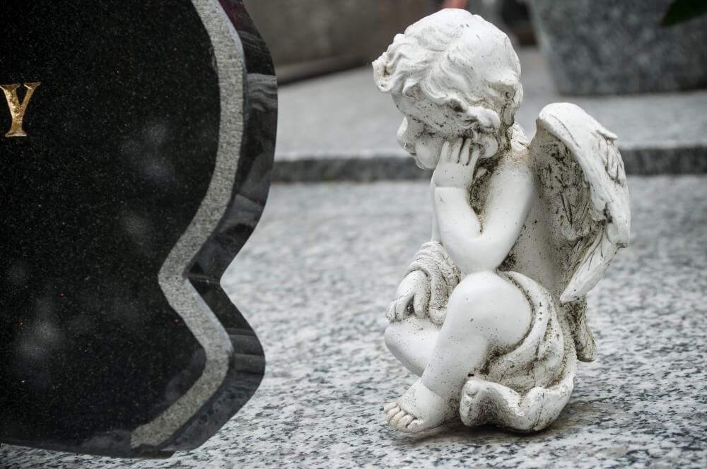 un bébé ange ailé en marbre blanc assis par terre devant une plaque mortuaire en marbre noir